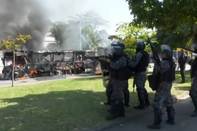 Foto mostra policais e um ônibus em chama aos fundos