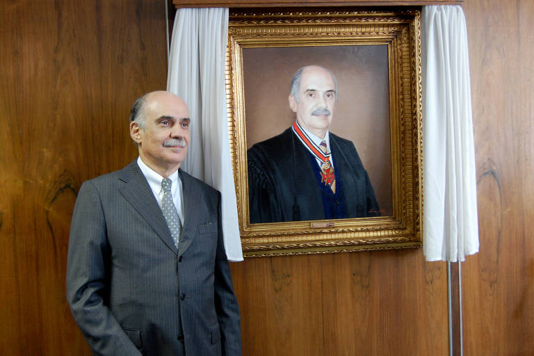 Homem vestido com terno e gravata, calvo e com bigode grisalho, ao lado de quadro com sua imagem em parede de madeira
