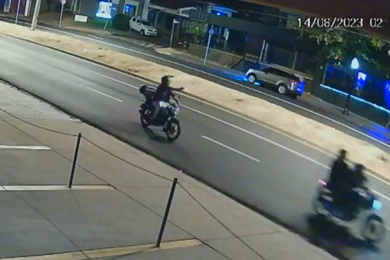 Um homem em uma motocicleta na pista está com uma das mãos esticadas, segurando uma arma, apontada em direção a outra motocicleta. A moto que tenta fugir tem dois ocupantes. Do outro lado da avenida, um veículo cinza em sentido oposto