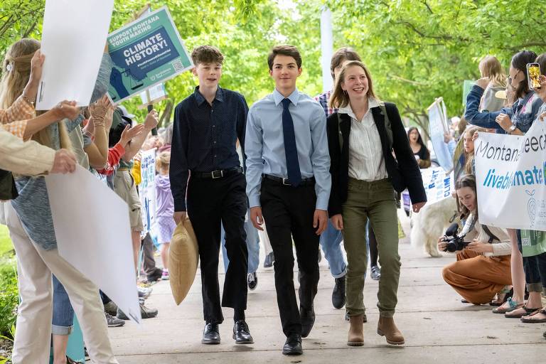 Adolescentes vestidos com roupas formais sorriem e caminham por uma rua, enquanto outros jovens seguram cartazes e bandeiras em apoio a eles