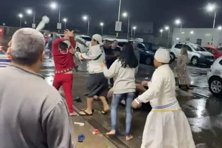 três pessoas brigam em estacionamento de supermercado observadas por mulher de turbante e saia branca e por homem de moletom