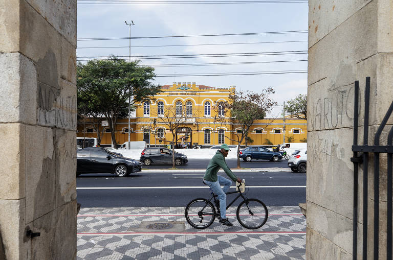 Ciclista passa pela ciclovia em cima da calçada. Em frente, carros passam pela avenida e ao fundo o prédio amarelo do Batalhão Tobias Aguiar