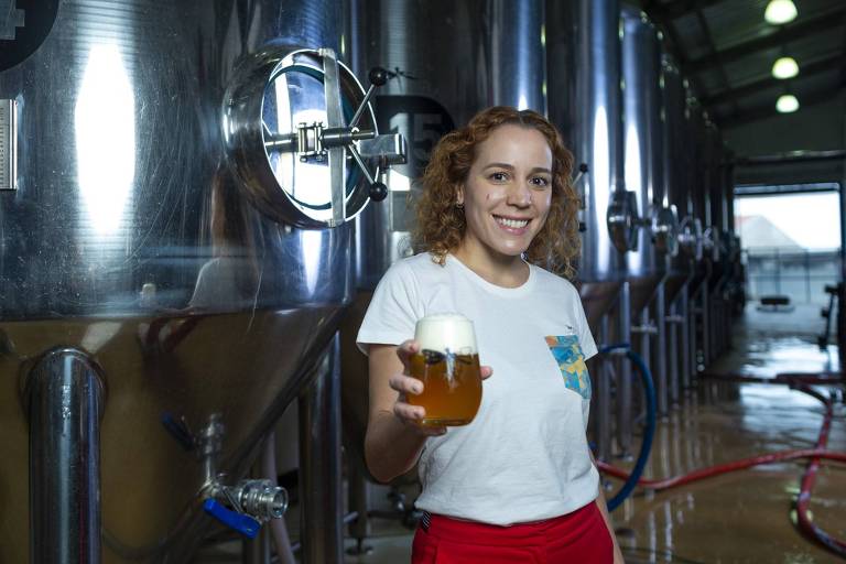 A empresária Luiza Tolosa aparece em sua cervejaria segurando um copo de cerveja com cilindros metálicos da bebida ao fundo. Ela veste blusa branca, calça vermelha e tem os cabelos loiros e cacheados