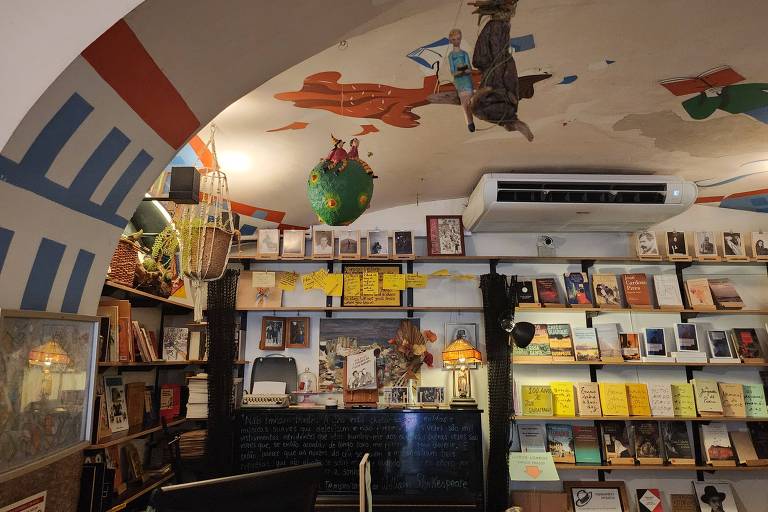 Foto da livraria Menina e Moça em Lisboa mostra prateleiras com livros, um piano ao fundo com objetos diferentes e utensílios de decoração diferente nas paredes e no teto