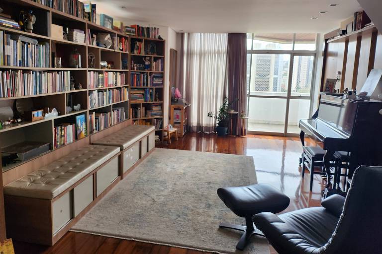 Imagem da biblioteca da Cynthia, com um tapete no meio, livros ao lado esquerdo em prateleiras sob dois bancos, um piano e uma poltrona ao lado direito