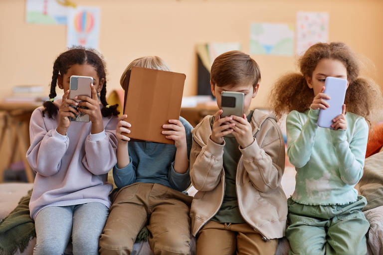 Crianças olham fixamente para telas de celular, que cobrem seus rostos
