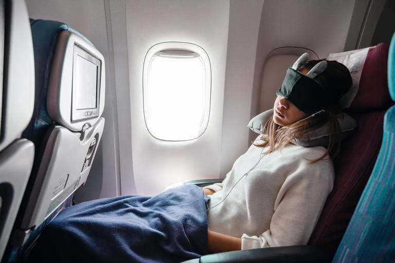 Fotografia colorida mostra um passageiro branco de cabelos longos e claros viajando em uma poltrona ao lado da janela do avião; a pessoa está com uma máscara de dormir, uma camisa branca e coberta com uma manta azul marinho