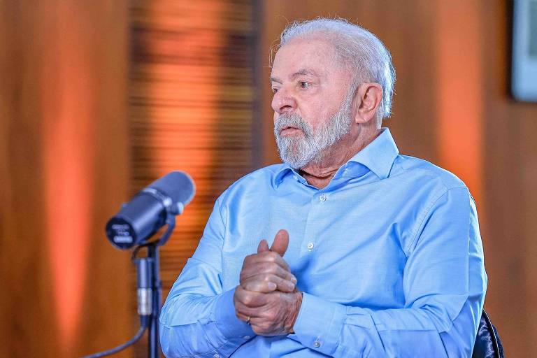 O presidente Luiz Inácio Lula da Silva aparece sentado em frente a um microfone em uma sala com pareces de acabamento amadeirado; usando camisa social de cor azul-claro, ele mantém expressão facial séria e aperta as mãos