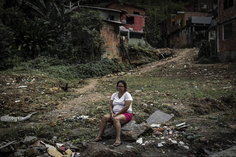 Mulher sentada sobre pedaços de casa, numa área aberta, com casas parcialmente destruídas ao fundo