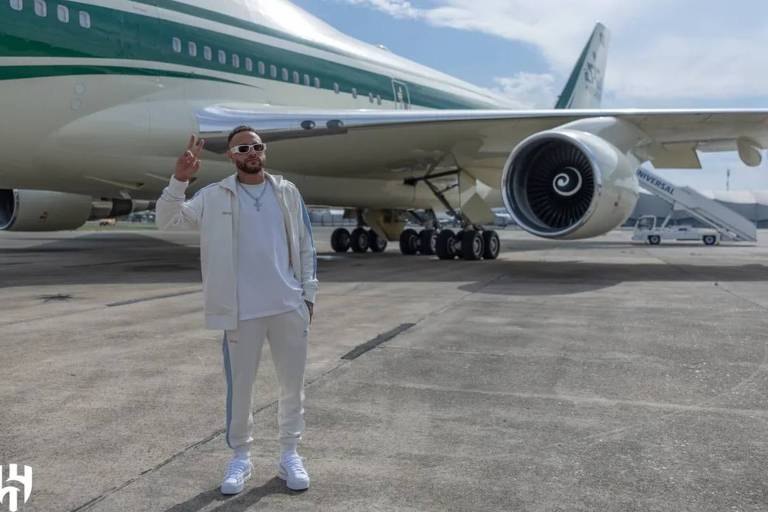 Em foto colorida, homem vestido com um jogging branco acena na entrada de um avião