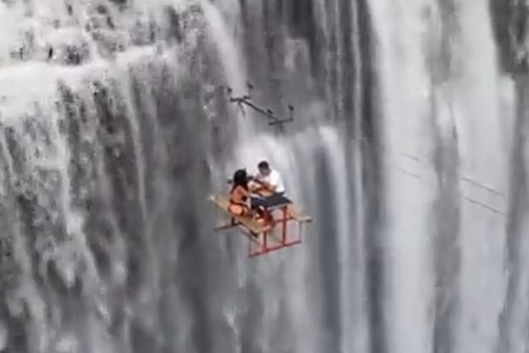 Em foto colorida, um casal faz piquenique suspenso em uma cachoeira