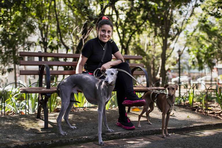 Fotografia colorida mostra Thais sentada no banco de uma praça segurando dois cães pela coleira; ela é uma mulher branca de cabelos castanhos e sorri para a foto