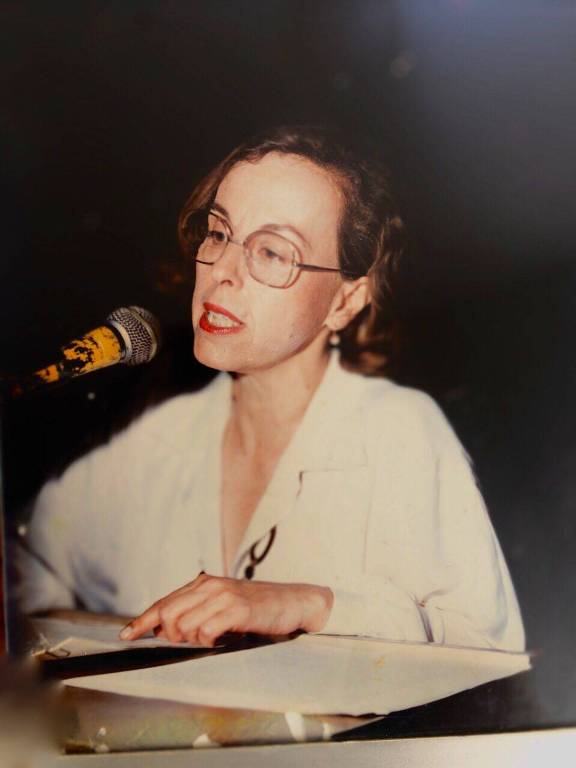 fotografia antiga mostra a professora homenageada falando em um microfone, com os braços apoiados sobre mesa