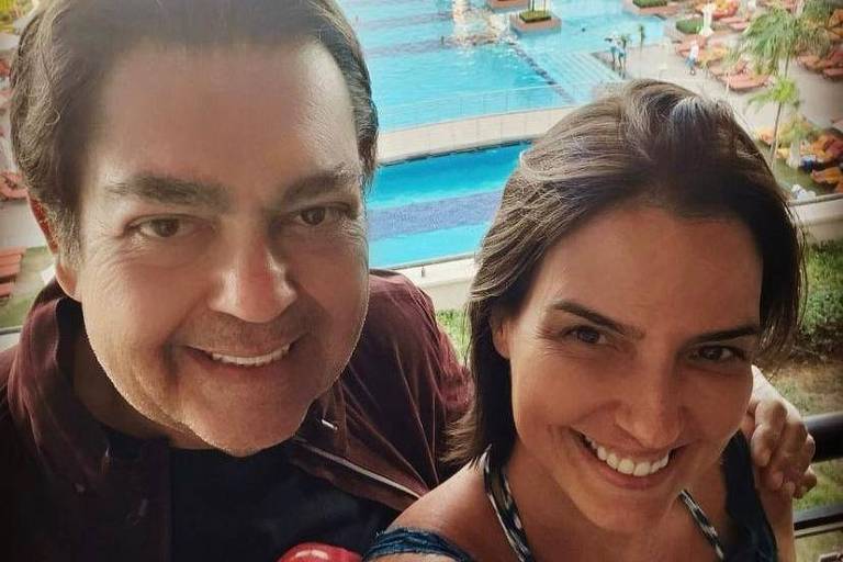 Fausto Silva e a mulher, morena de cabelos lisos, aparecem numa selfie, do ombro para cima,tendo ao fundo uma grande piscina 