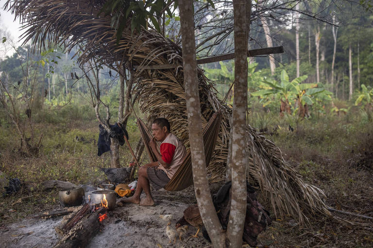 Homem indígena, usando camiseta branca com as mangas vermlhas, sentado no chão sob abrigo de folhas de palmeira, ao lado de fogueira
