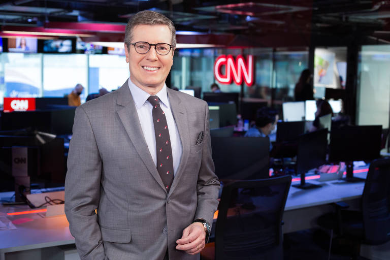 O jornalista Márcio Gomes posa para foto no estúdio da CNN em São Paulo; ele é um homem branco, usa óculos e veste um terno cinza e uma gravata cinza escura com detalhes em vermelho