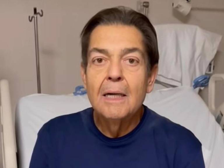 O apresentador Fausto SIlva está sentado numa cama de hospital, com camisa azul marinho