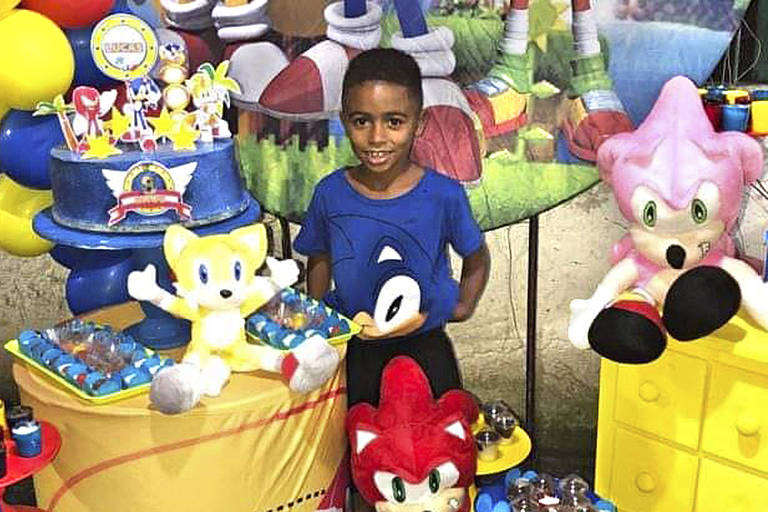 Na imagem, Lucas aparece comemorando seu aniversário em uma casa de festa infantil. Ele sorri para a câmera e posa entre brinquedos de pelúcia do desenho Sonic