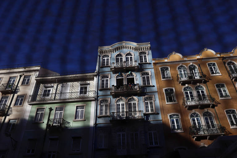 Quatro prédios prédios residenciais em Lisboa, cidade onde o preço dos imóveis disparou nos últimos cinco anos, vistos através de uma grade. O céu está azul e os edifícios são coloridos. 