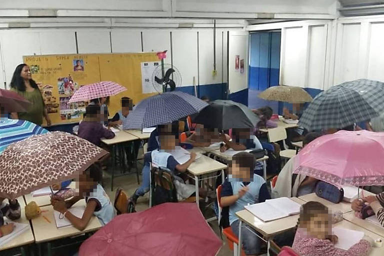 Crianças, com rosto borrado, sentadas em carteiras. Elas seguram sobre suas cabeças guarda-chuvas de cores diversas. Há um guarda-chuva rosa, outro em tons de azul, outro em tons de marrom e um outro vermelho. No fundo, uma mulher, professora, em pé, olhando em direção às crianças. As paredes da sala de aula são pintadas em azul e branco