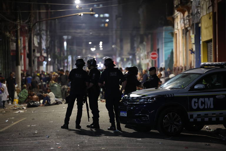 foto mostra agentes de costas em rua à noite. do lado direito da imagem, uma viatura em que se pode ler GCM, sigla da guarda. ao fundo, multidão 