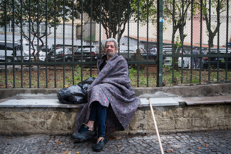 Morador de rua envolto em um cobertor e sentado numa mureta à frente de uma grade