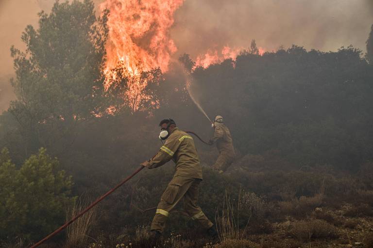 Dois bombeiros usam mangueiras contra fogo; há chamas altas na vegetação e muita fumaça 