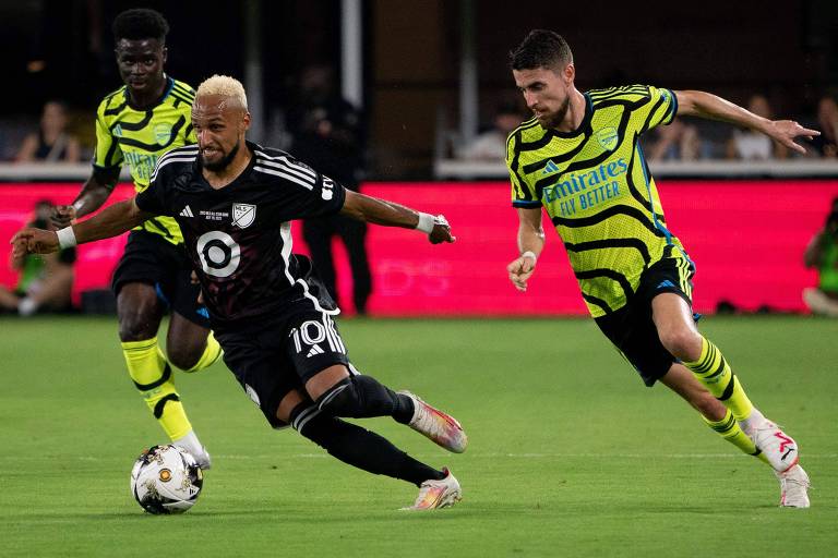 Imagem mostra três jogadores de futebol correndo atrás de bola em um campo.