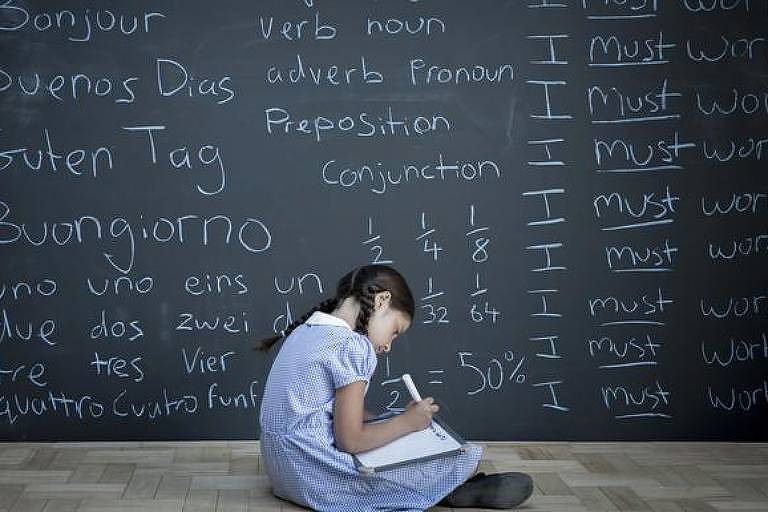 Menina escrevendo em caderno em frente a lousa com palavras em várias línguas
