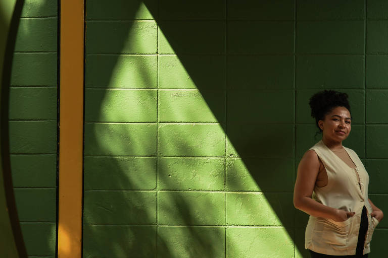 Uma mulher negra, com um coque, sorri discretamente. Ela usa jaleco cor creme e ao fundo há uma parede verde onde bate o reflexo da luz do sol em uma parte