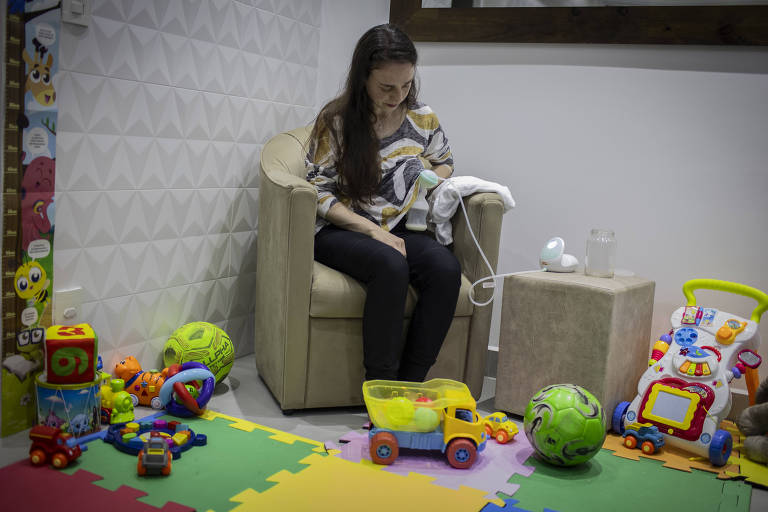 Mulher jovem está sentada numa cadeira, em meio a brinquedos de um bebê, retirando leite do peito