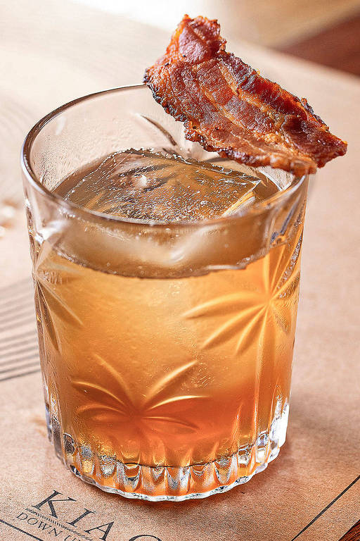 A foto mostra o Down Under, drinque do Kia Ora servido em um copo baixo com um gelo grande e sobre a borda do copo, uma fatia de bacon crocante
