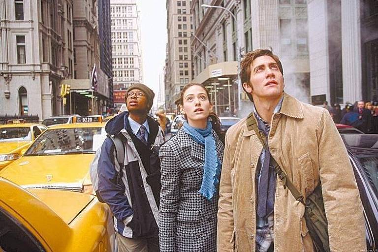 Um jovem negro (à esq.), uma jovem branca e um rapaz branco vestem roupas de inverno e estão parados no meio de uma rua em Nova York. Ao redor deles, carros e táxis amarelos parados. Eles olham para cima com expressão de surpresa e choque