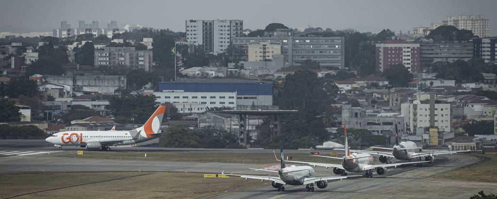 Imagem mostra fila de aviões em pista de aeroporto. No fundo, é possível ver vários prédios.