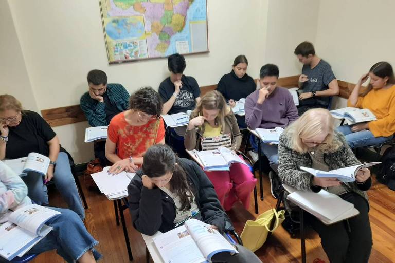 Cerca de 10 pessoas sentadas lendo livros em sala de aula, com mapa do Brasil atrás