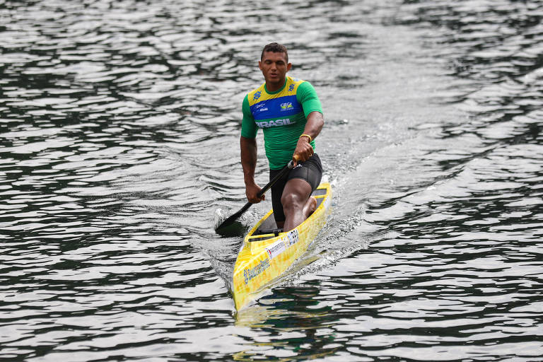 Um homem pardo, vestindo uniforme do time brasileiro, rema ajoelhado numa canoa