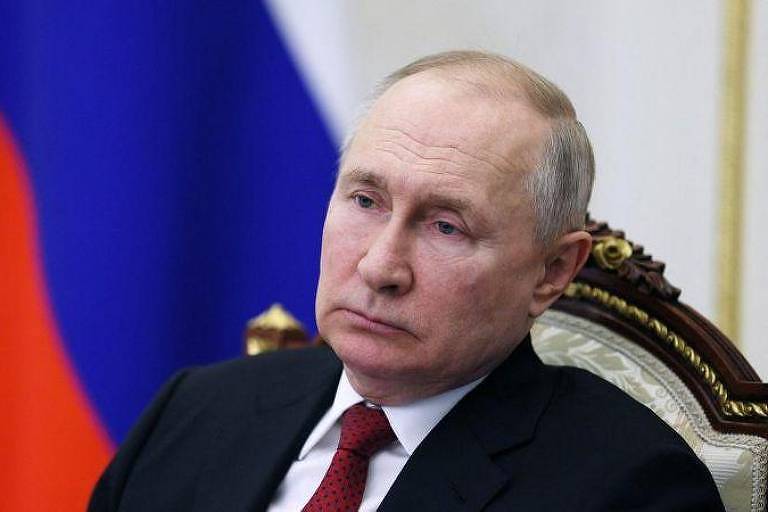 Retrato do presidente russo Vladimir Putin; ele é um homem branco e calvo com cabelos grisalhos; ele veste termo preto com camisa branca e gravata vermelha; ao fundo, está hasteada uma bandeira da Rússia (formada por três faixas horizontais nas cores branca, azul e vermelha)