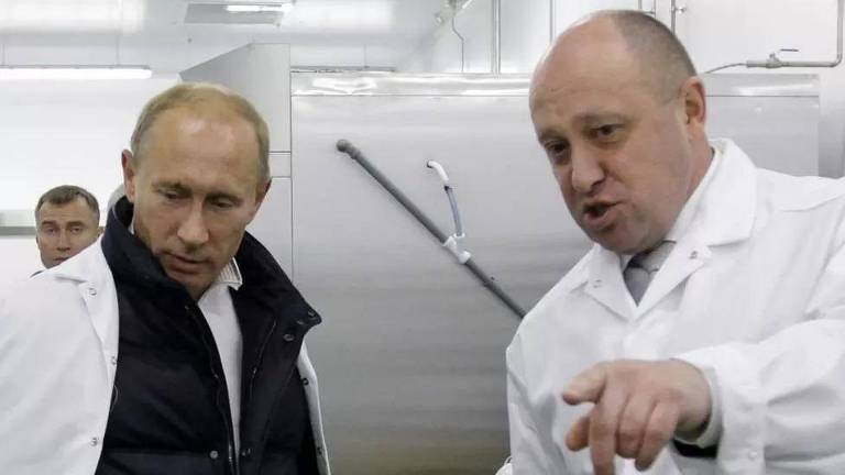 Retrato do presidente russo Vladimir Putin ao lado de Ievguêni Prigojin, líder do Grupo Wagner; ambos são homens brancos e calvos, vestidos com jalecos brancos em um laboratório; Putin está à esquerda e, além do jaleco, usa colete preto