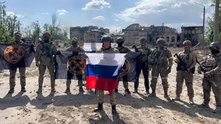 Foto mostra um grupo de homens com roupas militares; ao centro, um deles segura uma bandeira da Rússia (formada por três listras horizontais nas cores branca, azul e vermelha)