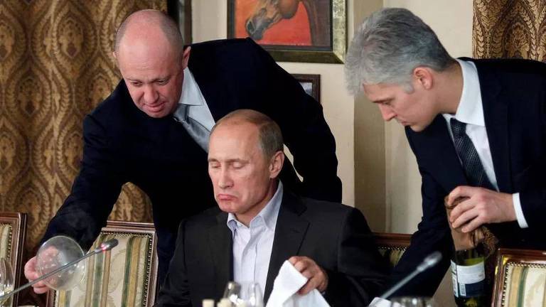 Foto mostra três homens: à esquerda, Prigojin serve um jantar a Putin, que está sentado à mesa, no centro da imagem; os três homens são brancos: Prigojin e Putin são calvos e têm cabelos grisalhos; o homem à direita está em pé e tem cabelos grisalhos; todos vestem ternos pretos