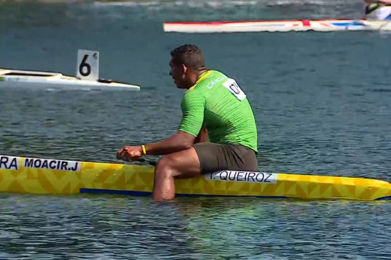 Isaquias, vestindo camisa verde e shorts preto, está sentado sobre a canoa, com os pés para fora mergulhados na água. Ele está com as costas curvadas 
