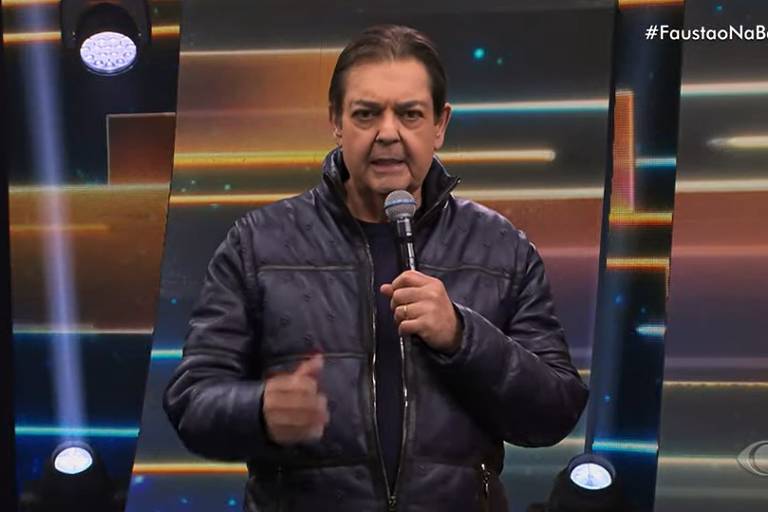 O apresentador Fausto Silva usa jaqueta preta e está em um local com luzes coloridas ao fundo 