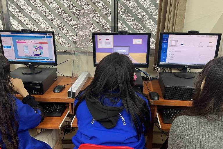 Três estudantes estão sentados em frente a computadores em uma sala de aula de informática, concentrados em suas telas, que exibem interfaces gráficas coloridas. Eles parecem estar engajados em uma atividade educacional, vestindo uniformes escolares azuis com detalhes em branco.