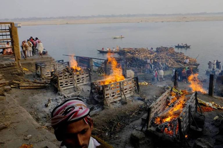 Imagem mostra pilhas de madeira em chamas, perto de rio, com algumas pessoas no entorno