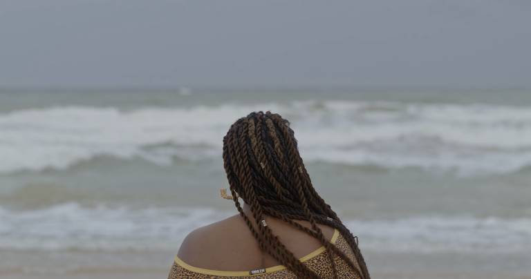 Mulher com dreads aparece de costas na foto, enquanto está em frente ao mar na praia