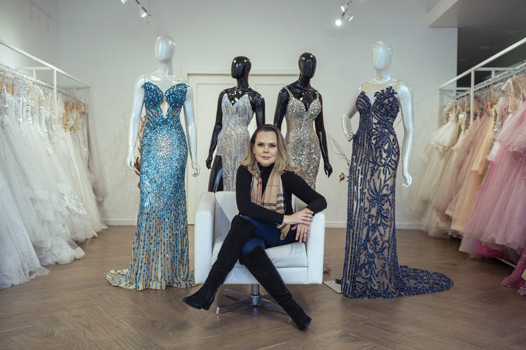 Daniela está sentada ao centro de seu ateliê, ao fundo aparecem quatro vestidos feitos por ela em manequins