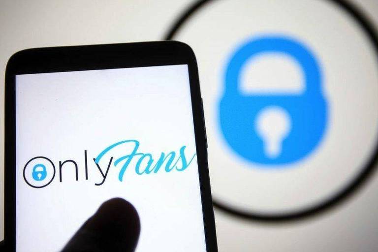Lucros do OnlyFans dispararam no ano passado. Pessoa acessa OnlyFans, num smartphone. Foto mostra apenas metade da tela e o dedo do usuário. Ao fundo há um painel com o logo da plataforma de conteúdo adulto.