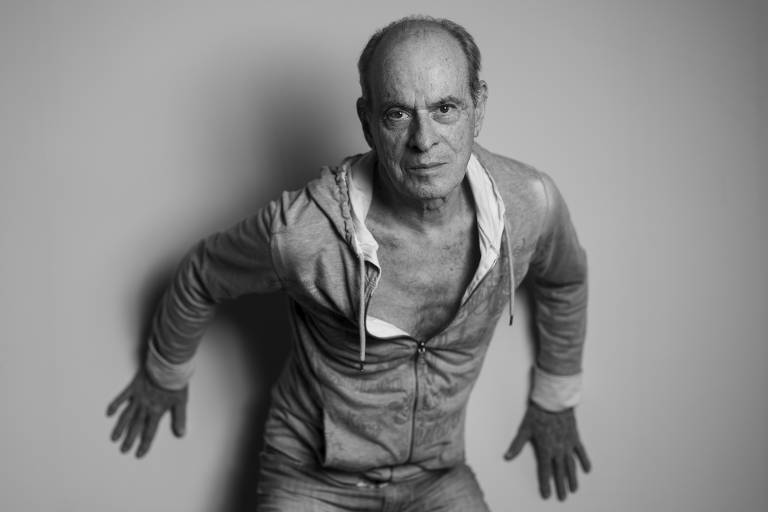 Imagem em preto e branco do cantor encostado na parede, com as mãos também apoiadas, olhando para a câmera. A camisa está aberta no peito até a metade