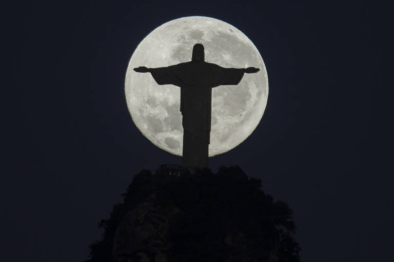 Silhueta da estátua do Cristo Redentor, de braços abertos. Ao fundo, a lua está cheia, redonda e clara. O céu está escuro.