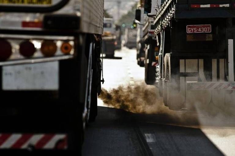Em avenida, foto dá foco à traseira de dois caminhões. Um deles despeja fumaça densa e de cor escura no asfalto